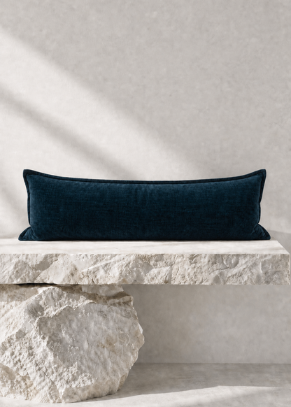 A dark navy blue velvet rectangular lumbar pillow displayed on a rustic white stone bench against a textured light gray wall with soft diagonal shadows.