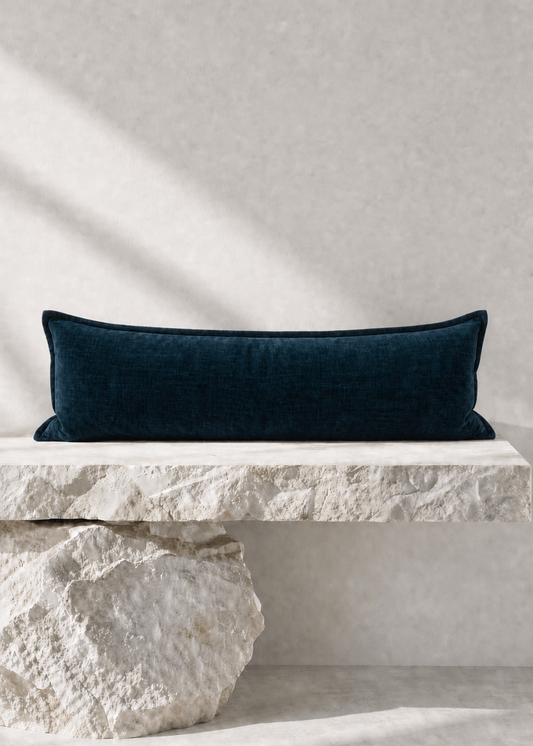 A dark navy blue velvet rectangular lumbar pillow displayed on a rustic white stone bench against a textured light gray wall with soft diagonal shadows.
