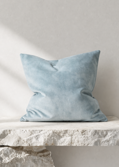 A square light blue velvet throw pillow resting on a textured white stone ledge against a neutral light gray wall.