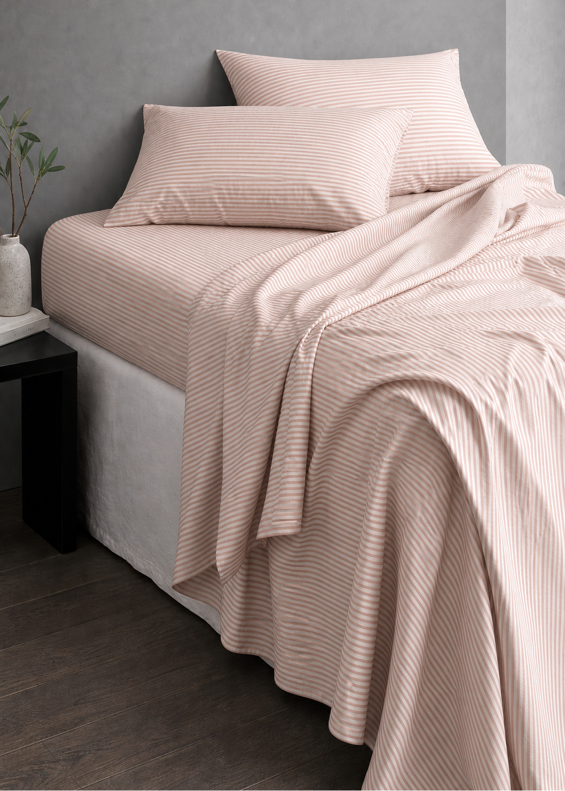 A bed dressed in pale blush pink striped bedding, including a flat sheet, pillowcases, and a draped blanket, set against a textured gray wall in a modern bedroom scene.