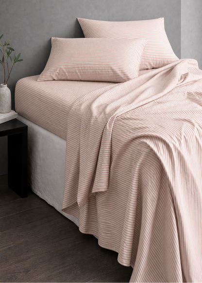 A bed dressed in pale blush pink striped bedding, including a flat sheet, pillowcases, and a draped blanket, set against a textured gray wall in a modern bedroom scene.
