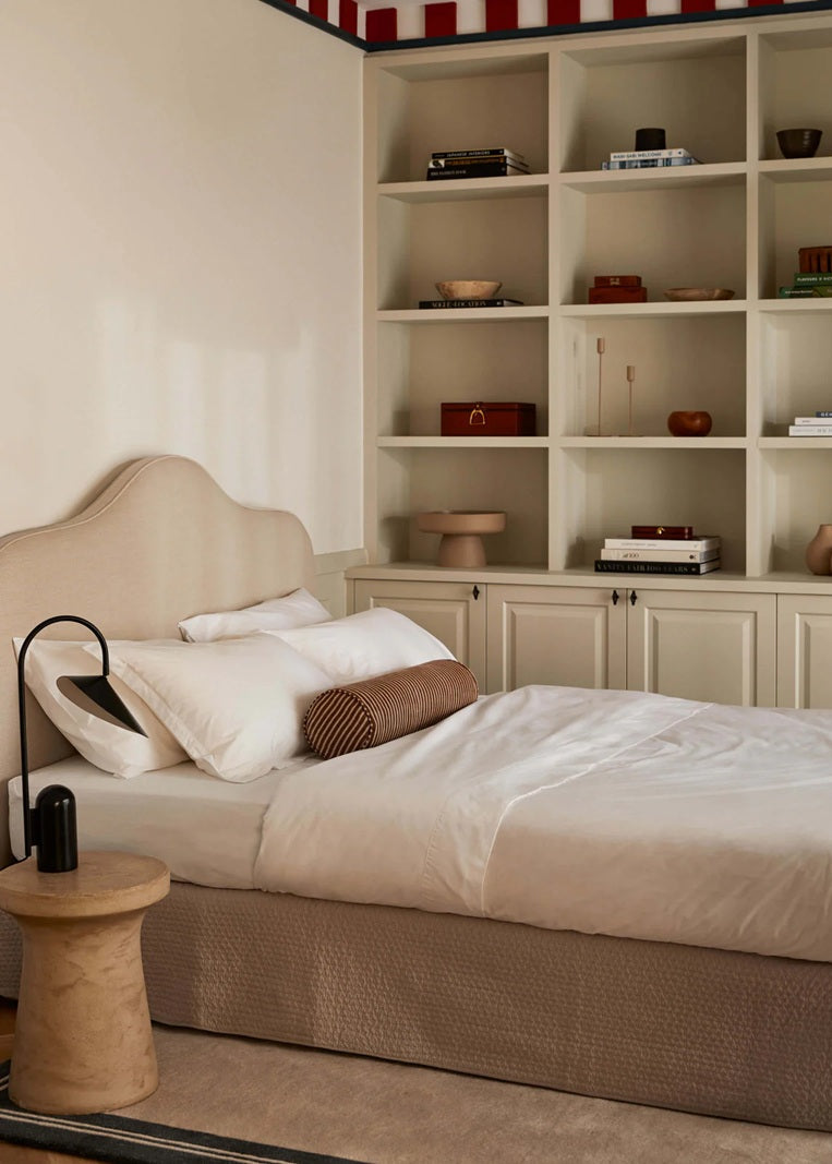 Neatly made bed with beige headboard in a bedroom with shelves displaying decorative items.