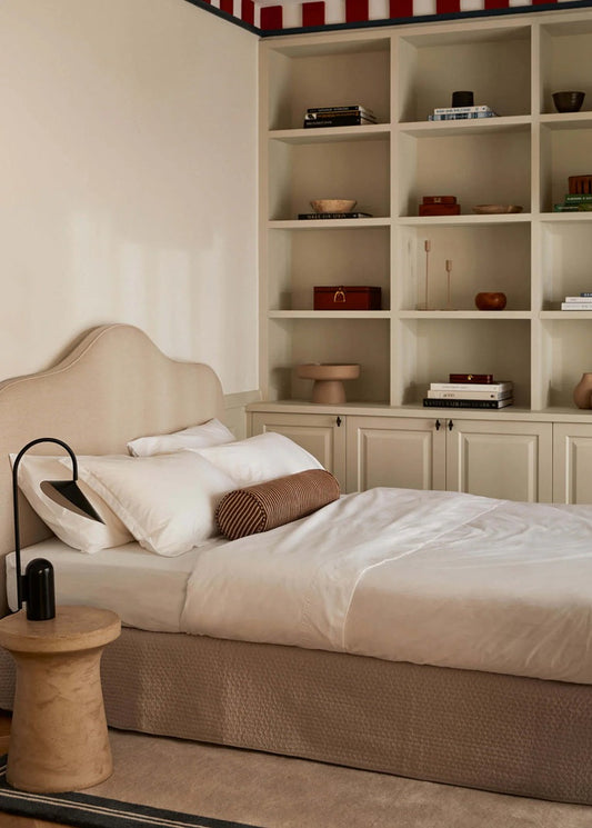 Neatly made bed with beige headboard in a bedroom with shelves displaying decorative items.