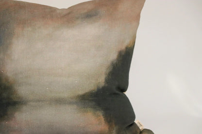 A close-up of a square throw pillow featuring a painterly, textured print with blended shades of gray, brown, beige, and dark charcoal.