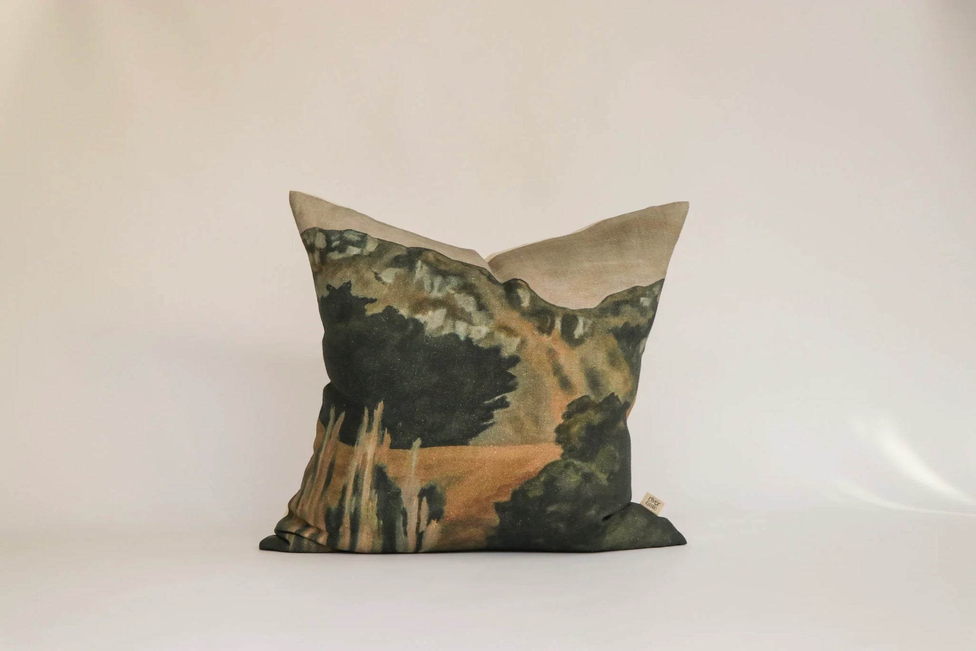 A square decorative throw pillow featuring a scenic landscape print with dark green trees, a winding beige path, and a light beige top border, set against a plain off-white background.