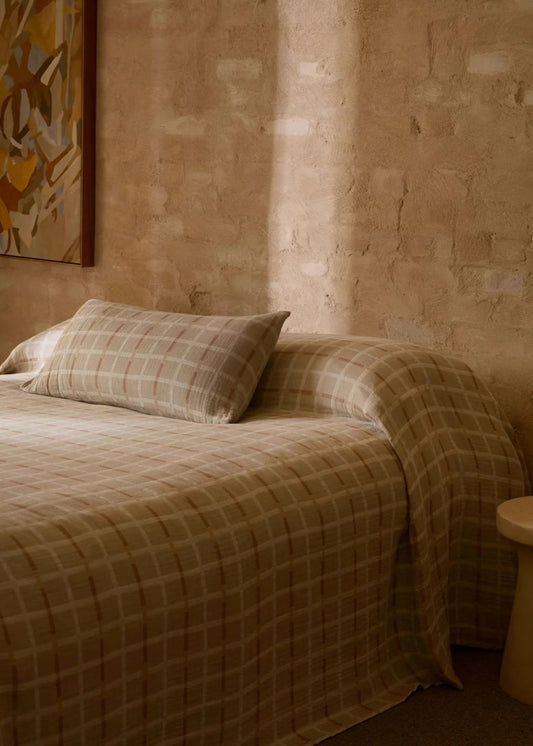 A bed dressed in beige and brown plaid bedding, set against a textured brick wall with warm sunlight casting shadows. A small round stool sits beside the bed, and a framed abstract painting hangs on the wall.