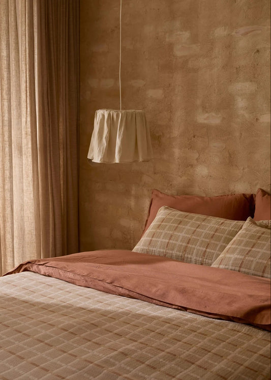 A cozy bedroom scene with a bed dressed in muted pink and beige plaid bedding, a textured warm brown plaster wall, beige curtains, and a hanging white lampshade.