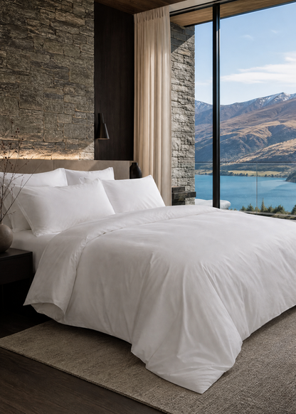 A luxury bedroom featuring a neatly made bed with crisp white bedding, set against a rustic stone wall and large floor-to-ceiling windows that offer a stunning view of a mountain lake.