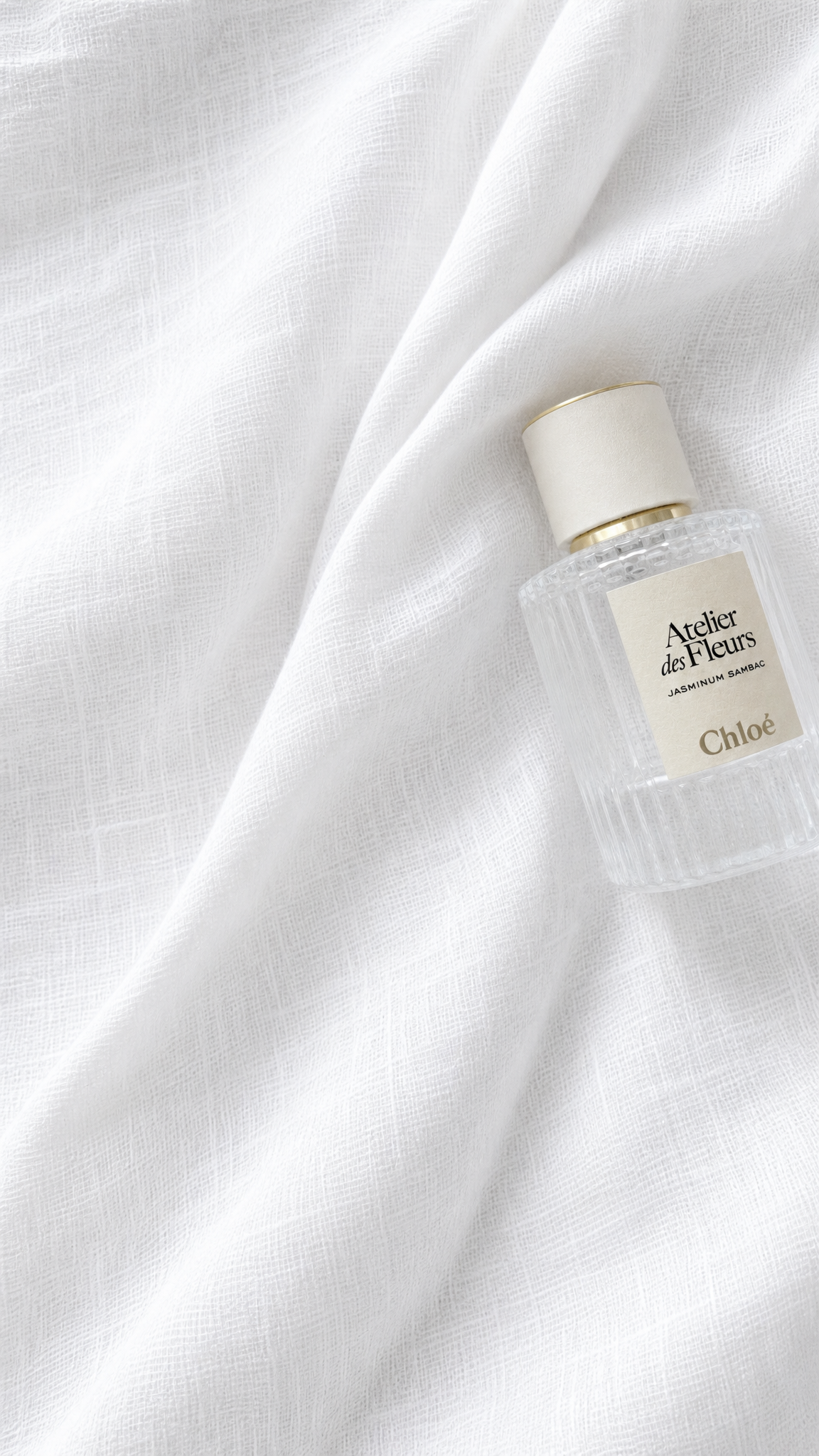 A small clear glass perfume bottle with a cream cap and cream label reading 'Arthor des Fleurs' and 'Chloé', placed on draped white fabric.