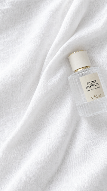 A small clear glass perfume bottle with a cream cap and cream label reading 'Arthor des Fleurs' and 'Chloé', placed on draped white fabric.