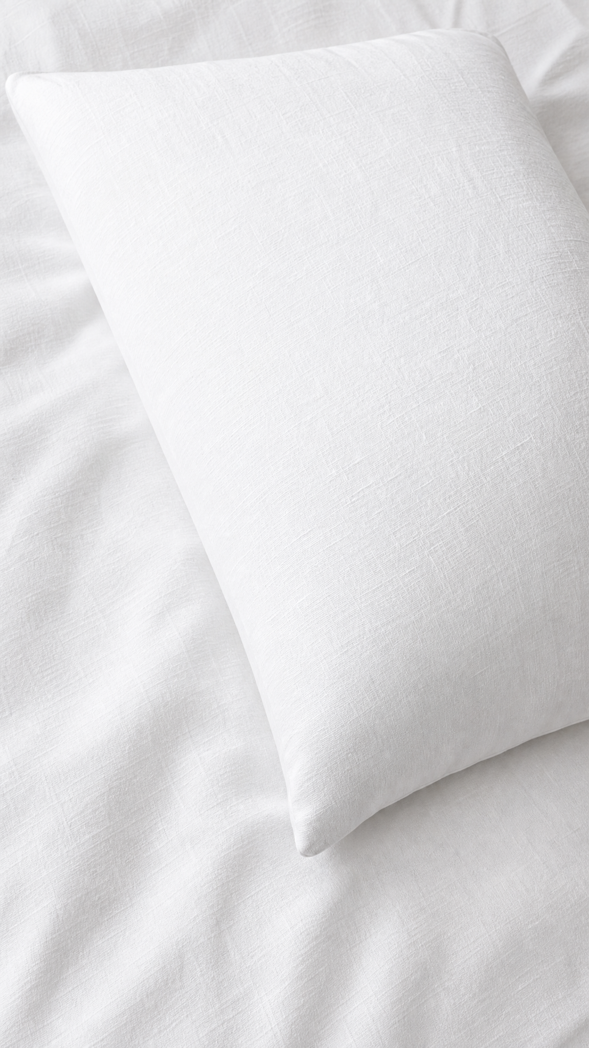 A white rectangular pillow resting on white bedding with a softly textured fabric surface.