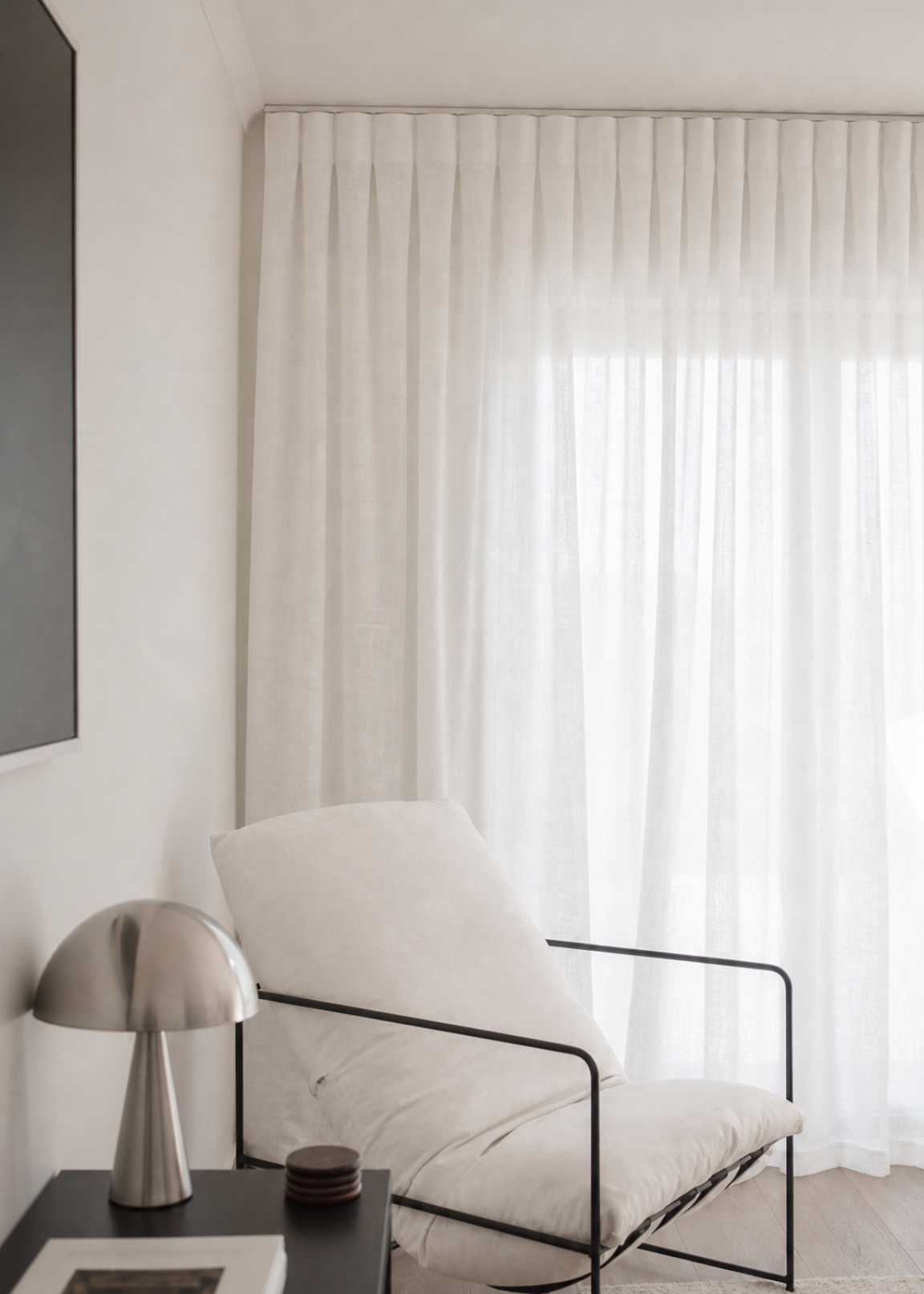 A modern white upholstered lounge chair with a black metal frame, positioned in a bright, minimalist room next to a black side table with a silver lamp and a framed print.