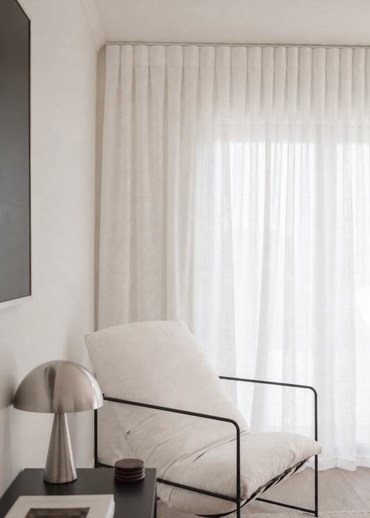A modern white upholstered lounge chair with a black metal frame, positioned in a bright, minimalist room next to a black side table with a silver lamp and a framed print.