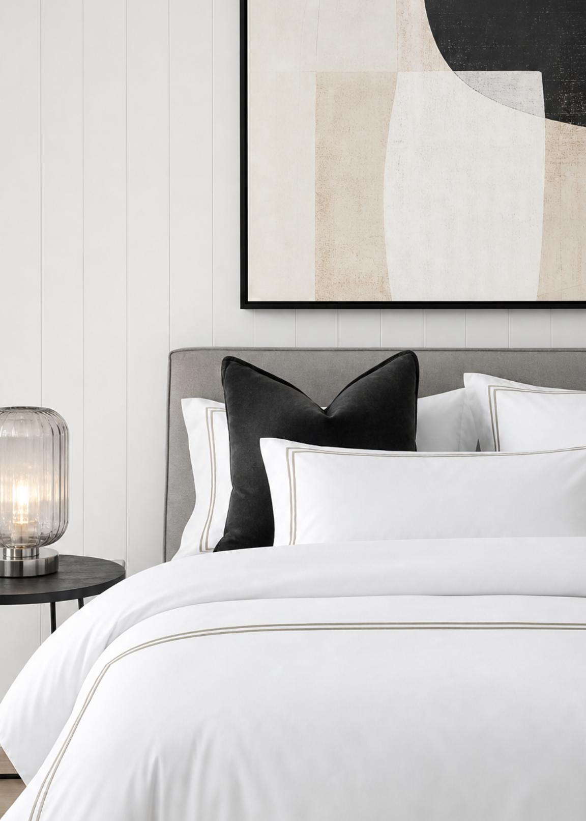 Neatly made bed with white bedding and black pillows, nightstand with lamp, and abstract art on wall.