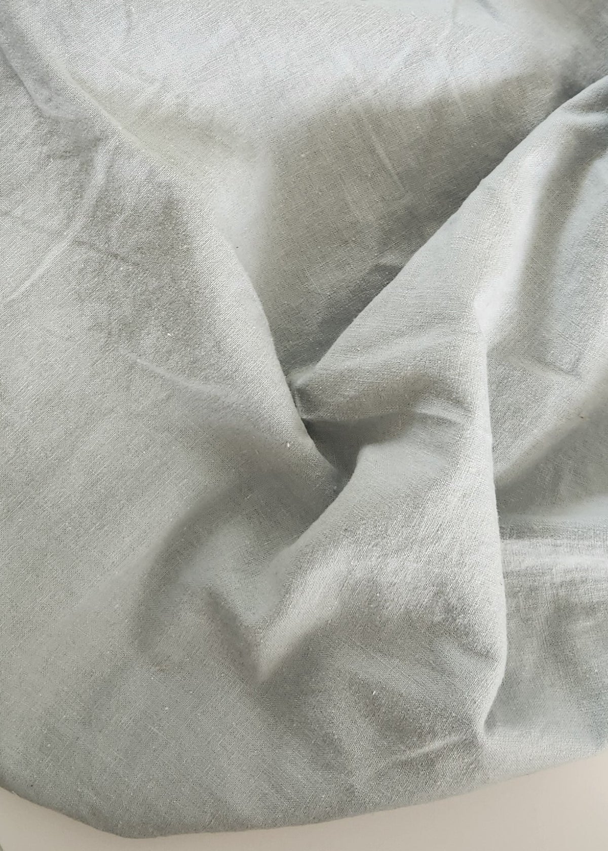 A light gray fabric bag or pouch made of a soft, slightly wrinkled material, draped over a white rectangular surface on a dark gray carpeted floor.