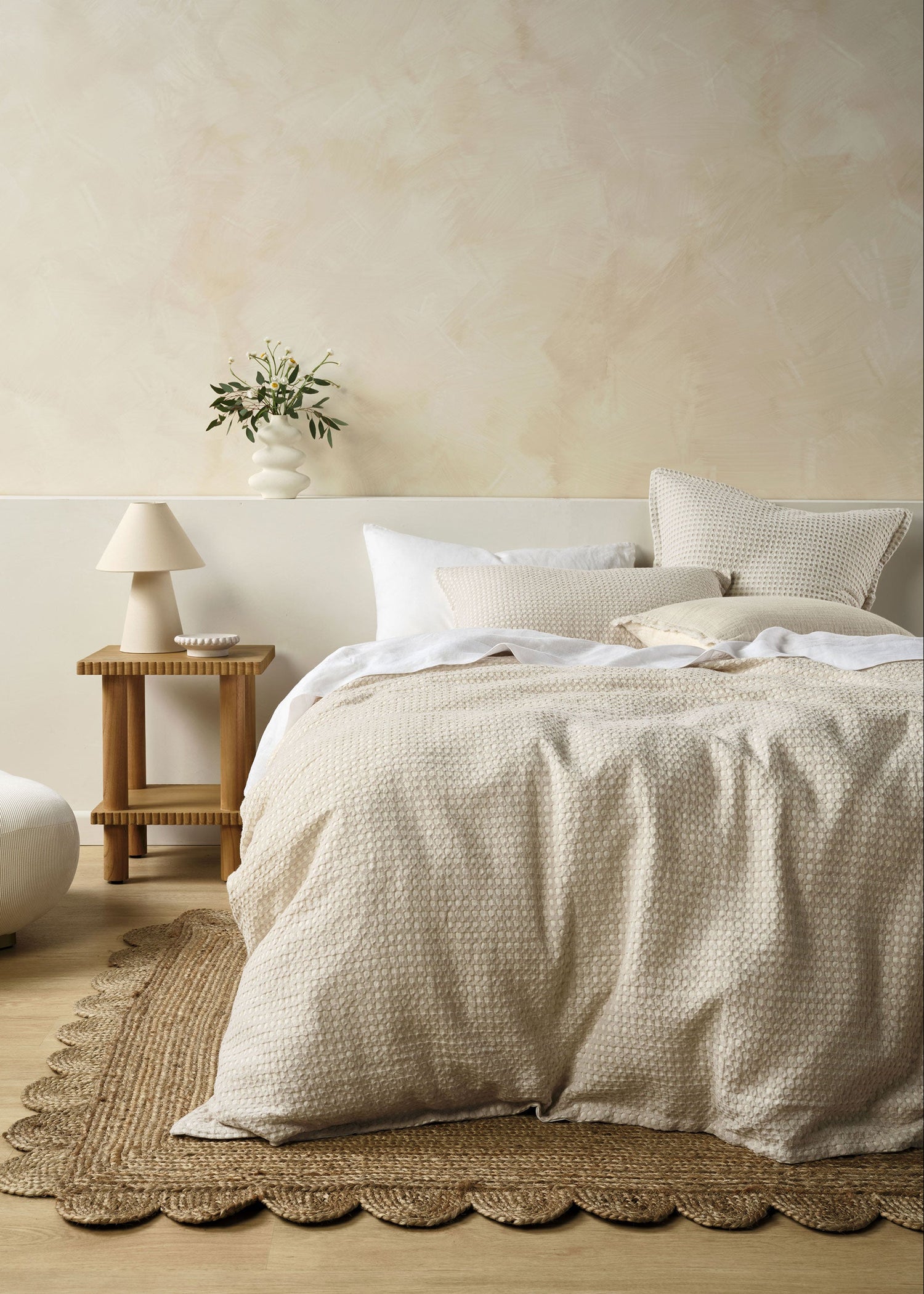 A bed with a beige Cotswold duvet cover set and matching pillowcases, presented in a bedroom setting with a wooden nightstand and lamp, and a plant in the background.