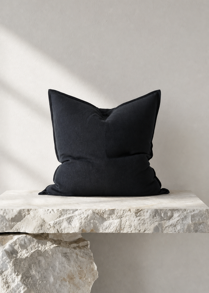 A black throw pillow with a slouchy, voluminous shape resting on a rustic white stone ledge against a textured light gray wall.