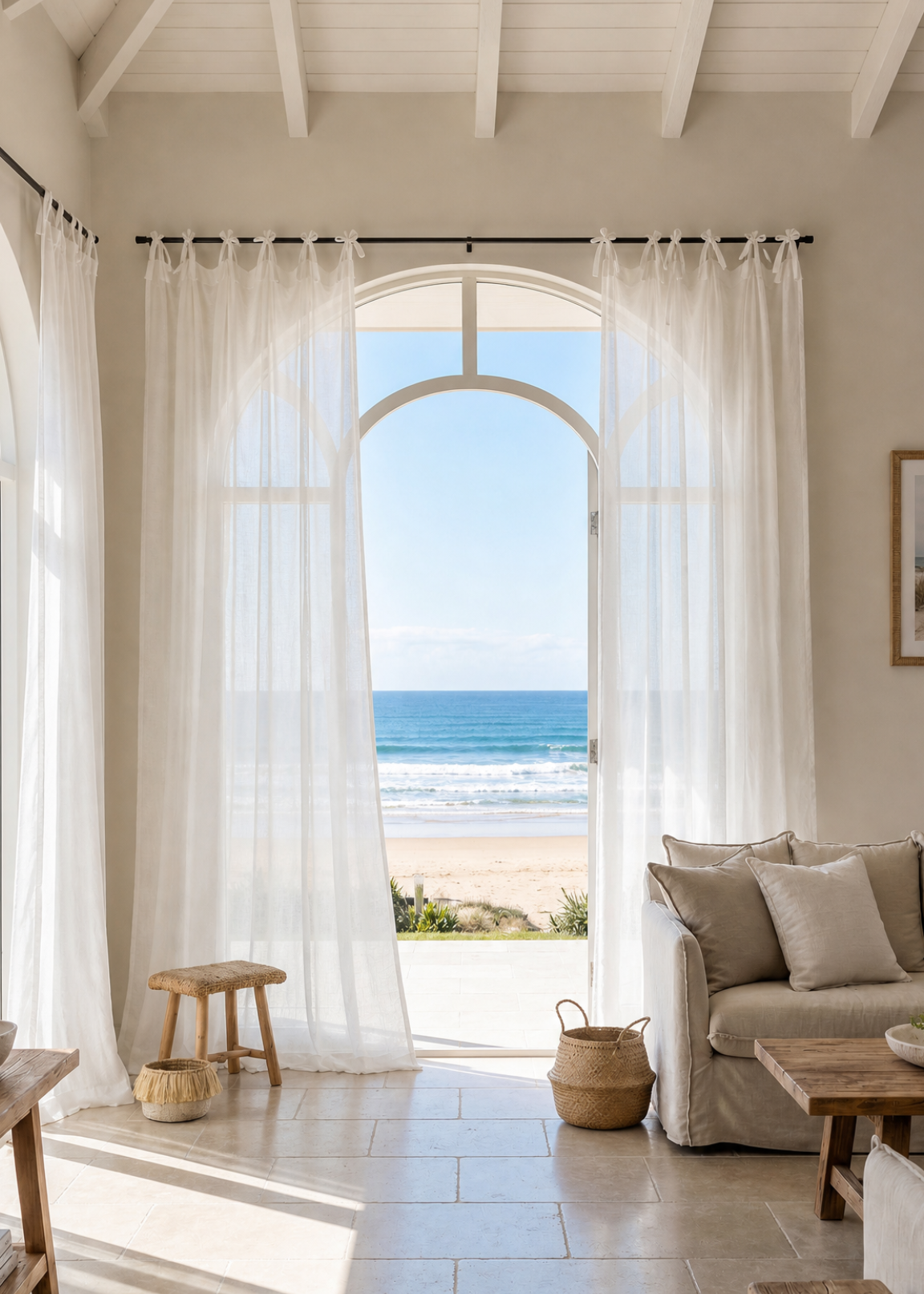A bright, modern living room with sheer white curtains framing a large arched window that looks out to a sandy beach and ocean. The room has light beige walls, a tiled floor, a gray sofa with pillows, and rustic wooden stools.