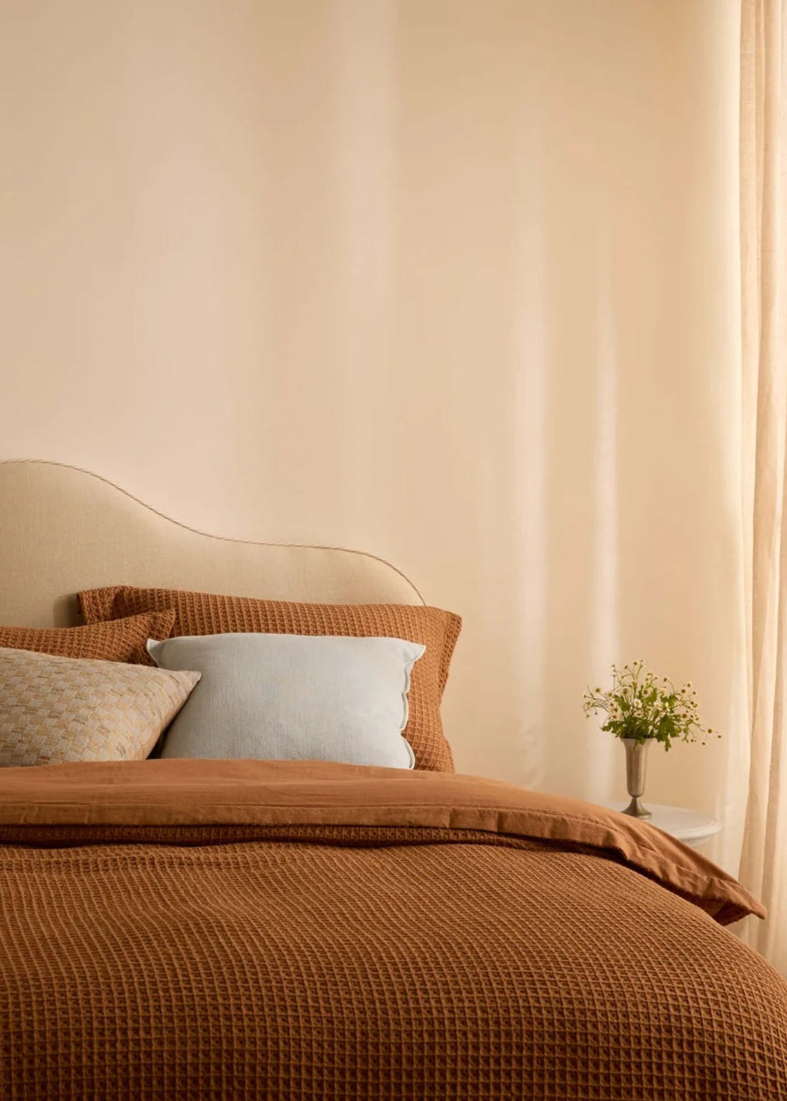 A cozy bedroom scene with a bed dressed in a textured rust-brown waffle blanket and matching pillow shams, accented by a light beige patterned pillow and a pale blue pillow. The room has warm beige walls, sheer cream curtains, and a small vase of flowers on a bedside table.