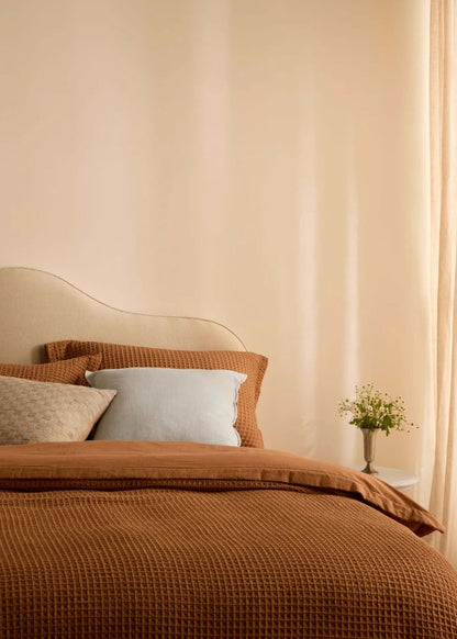 A cozy bedroom scene with a bed dressed in a textured rust-brown waffle blanket and matching pillow shams, accented by a light beige patterned pillow and a pale blue pillow. The room has warm beige walls, sheer cream curtains, and a small vase of flowers on a bedside table.