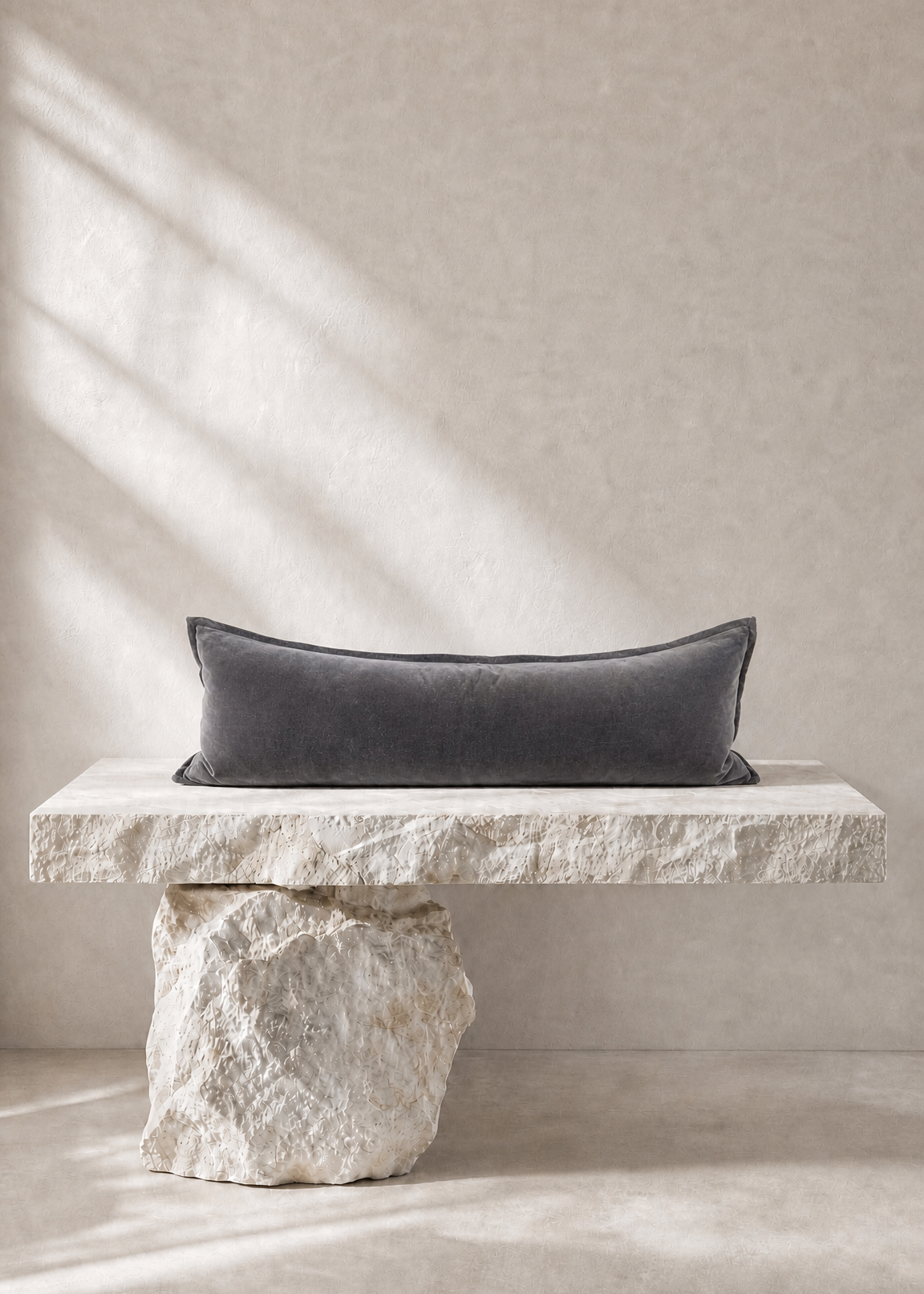 A dark charcoal gray rectangular lumbar pillow displayed on a rustic stone bench against a textured beige wall with soft window shadow light.