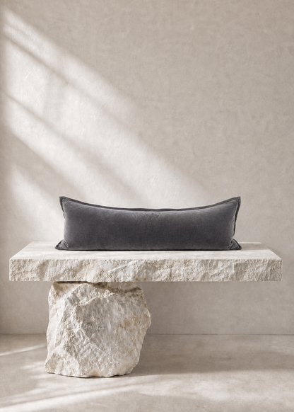 A dark charcoal gray rectangular lumbar pillow displayed on a rustic stone bench against a textured beige wall with soft window shadow light.