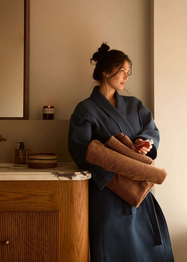 Woman in a blue robe holding folded towels in a bathroom.