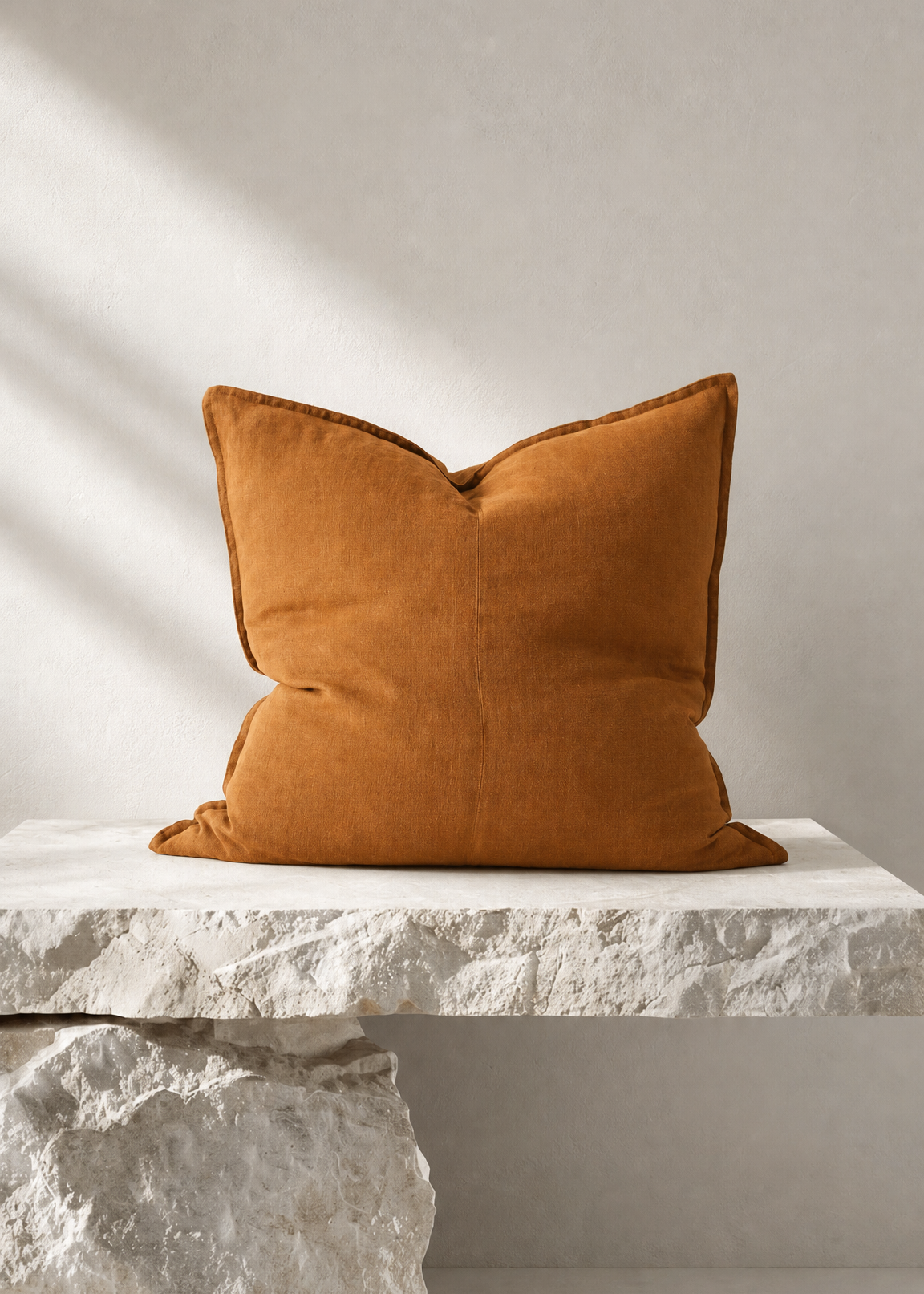 A square mustard-orange velvet throw pillow displayed on a white stone bench against a textured light gray wall with soft diagonal shadows.