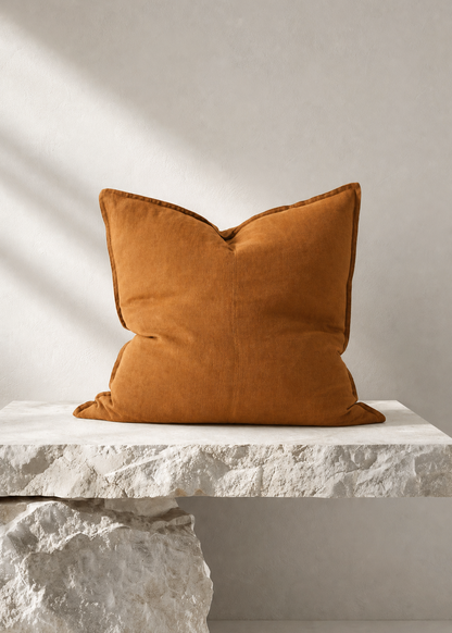 A square mustard-orange velvet throw pillow displayed on a white stone bench against a textured light gray wall with soft diagonal shadows.