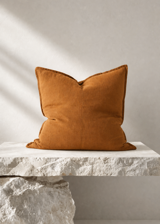 A square mustard-orange velvet throw pillow displayed on a white stone bench against a textured light gray wall with soft diagonal shadows.