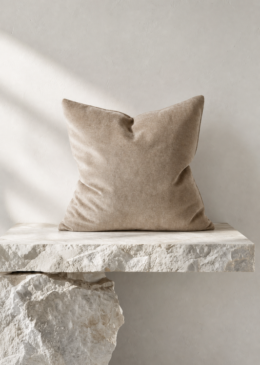 A square beige throw pillow with a textured linen-like fabric, resting on a rustic white stone bench against a neutral light gray wall.