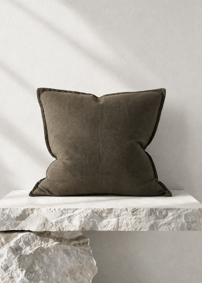 A square dark olive green throw pillow with piped edges, resting on a white stone bench against a textured white wall with soft diagonal shadows.