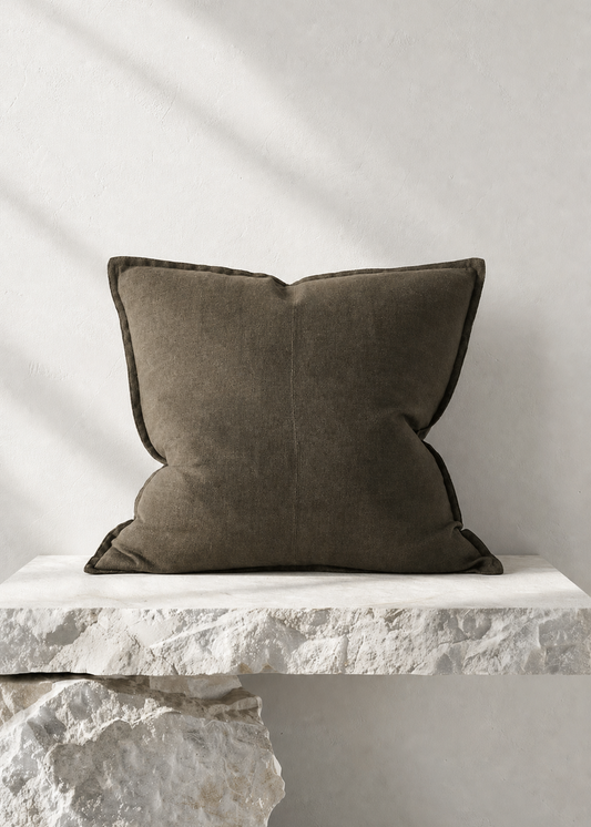 A square dark olive green throw pillow with piped edges, resting on a white stone bench against a textured white wall with soft diagonal shadows.