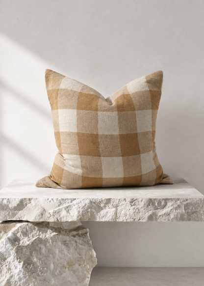 A square throw pillow with a tan and cream buffalo check plaid pattern, styled on a white stone bench against a neutral light gray wall.