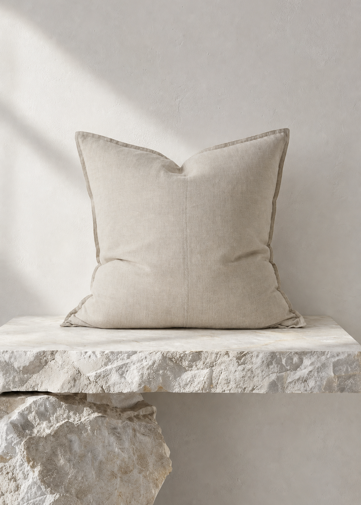 A square beige throw pillow with a subtle stitched center seam, resting on a rustic stone ledge against a textured light gray wall.