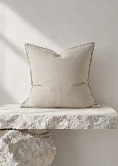 A square beige throw pillow with a subtle stitched center seam, resting on a rustic stone ledge against a textured light gray wall.