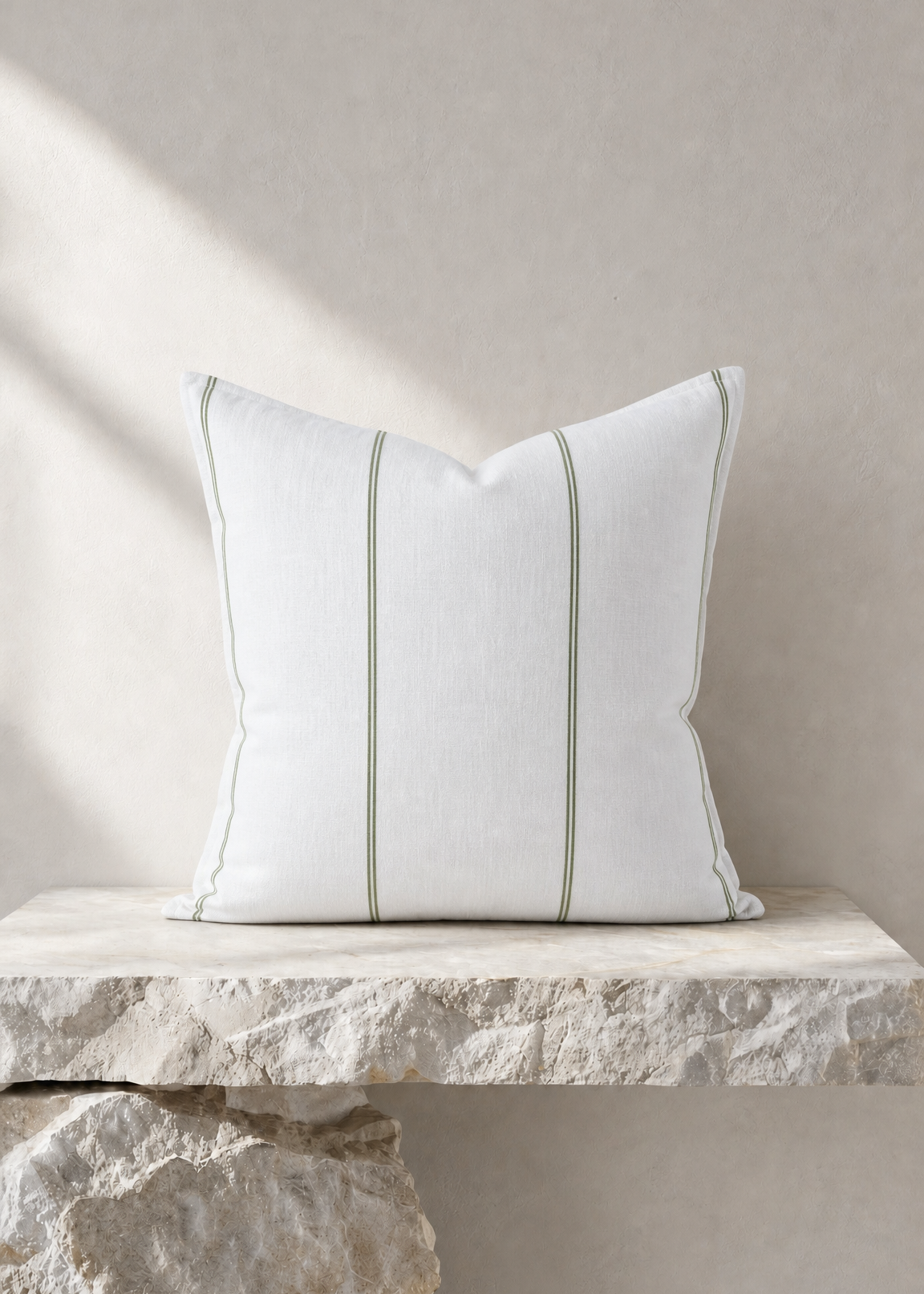 A square white throw pillow with two vertical light gray stripes and piped edges, resting on a rustic stone ledge against a textured beige wall.