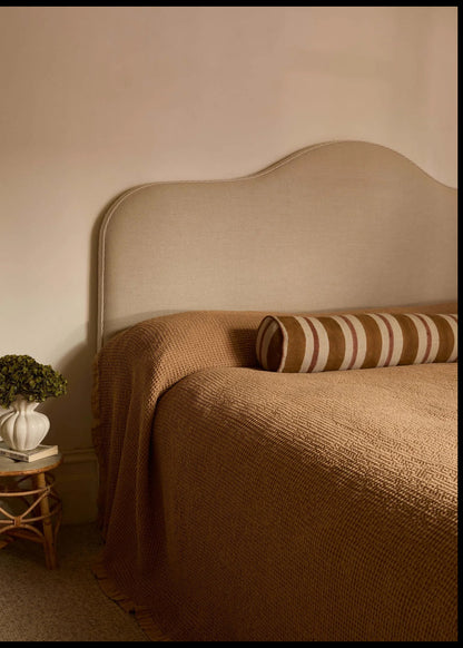 A cozy bedroom scene featuring a bed with a textured brown knit blanket, a matching scalloped upholstered headboard, and a striped brown and cream bolster pillow. A small wooden side table with a white vase of greenery sits to the left against a warm beige wall.