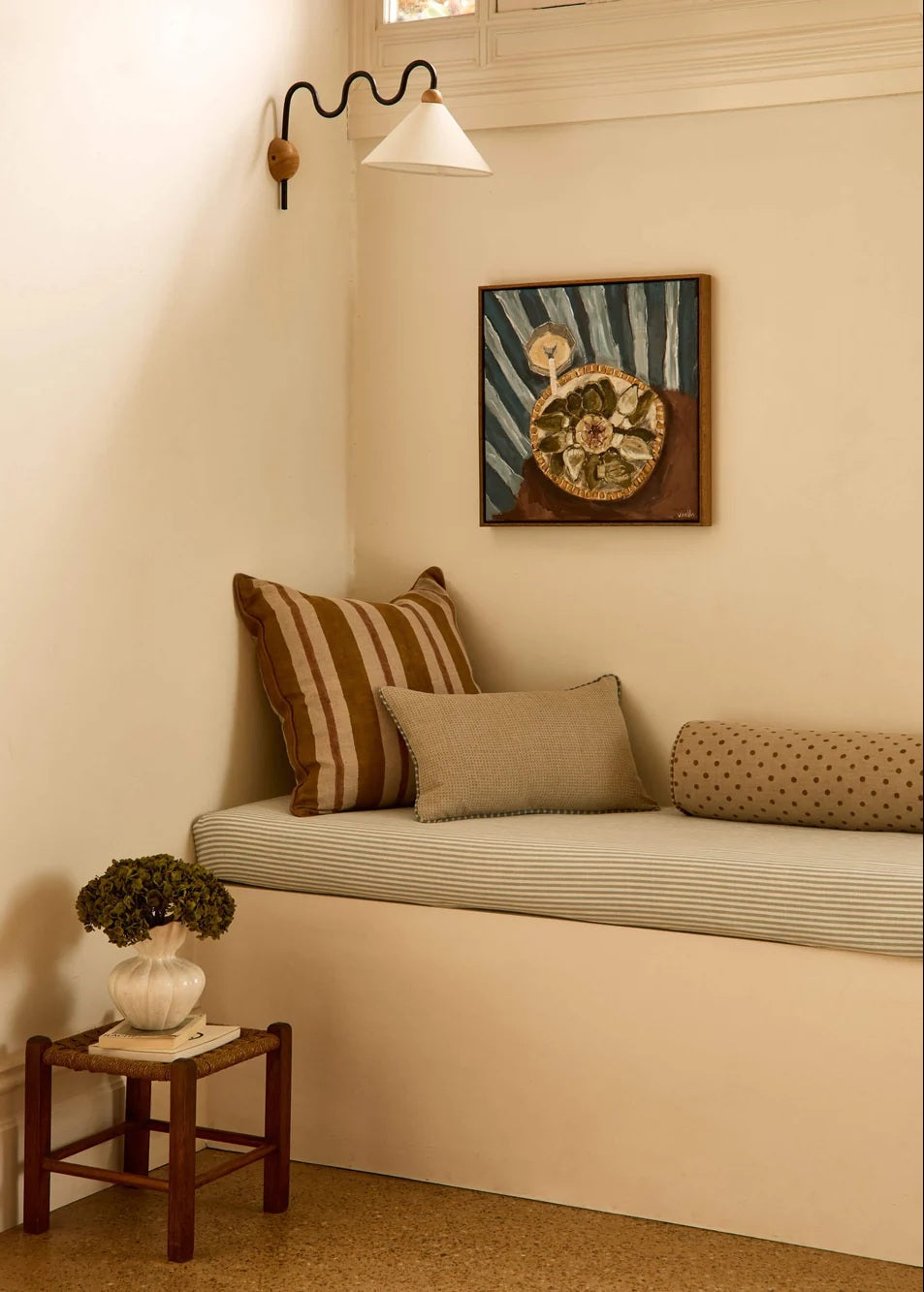 A cozy built-in window seat with cream and brown striped cushions, a small wooden side table with a potted plant, and a framed art print on the wall under a black wall sconce.