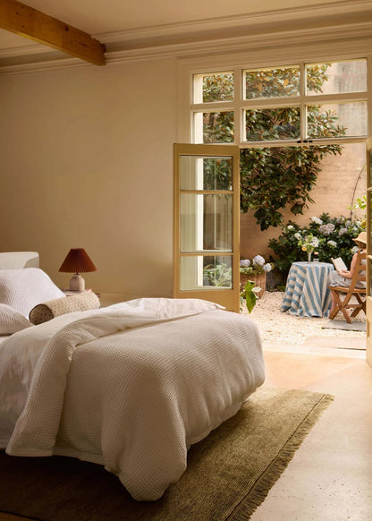 A beautifully styled bedroom with a bed dressed in white textured bedding, set in a warm neutral room with a wooden ceiling beam. Large open French doors look out onto a small garden patio with a table covered in a blue and white striped cloth.
