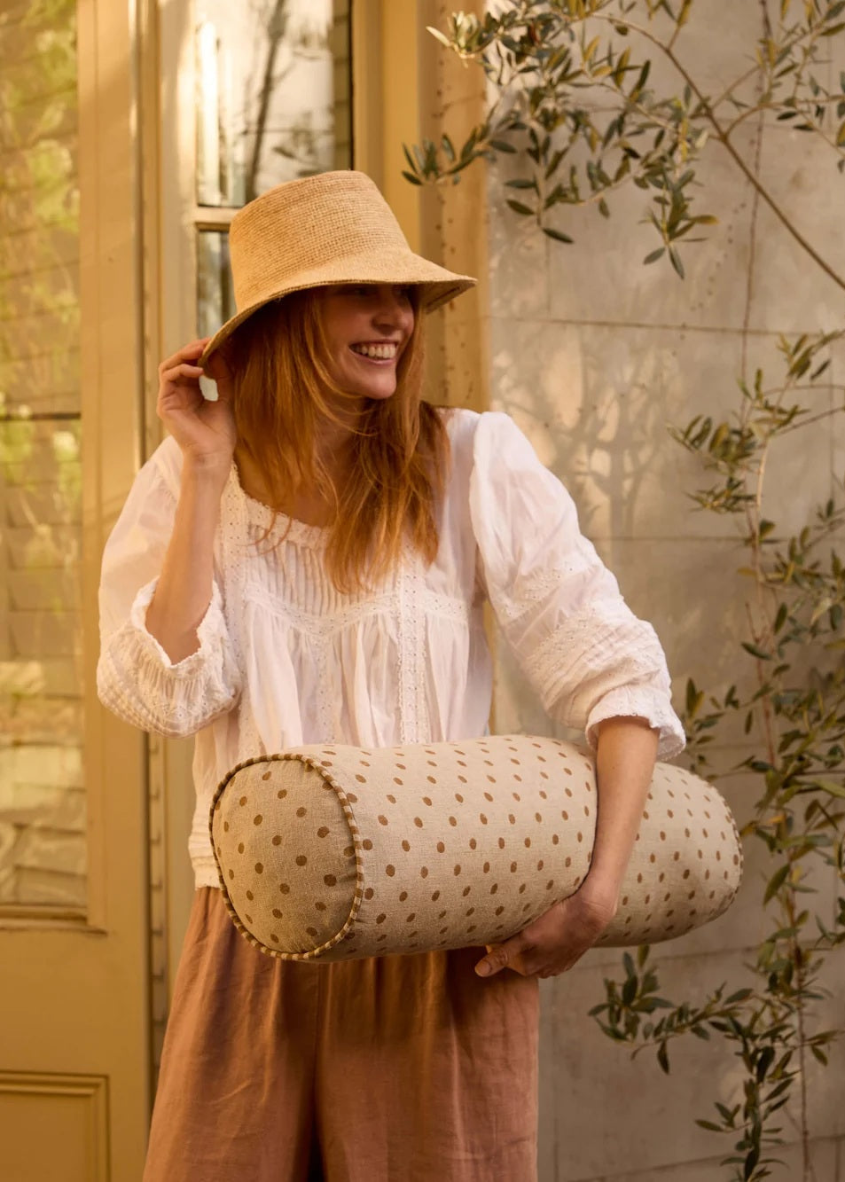 A woman in a bohemian-style outfit with a straw hat and white blouse holds a cylindrical bolster pillow with a beige and brown polka dot pattern in a warm, sunlit room.
