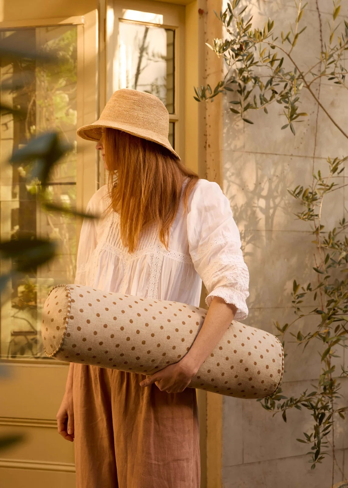 A woman in a white blouse, brown skirt, and straw hat carries a cylindrical lumbar pillow with a beige and brown polka dot pattern in a warm, sunlit room.