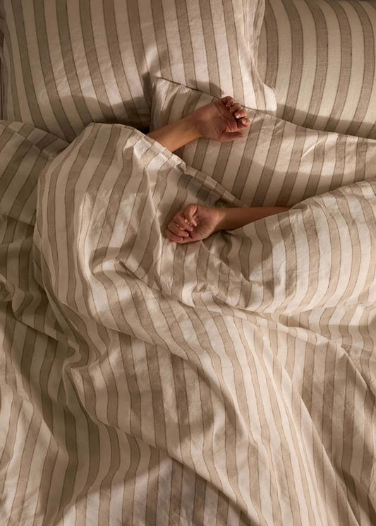 Person lying in bed under striped sheets with sunlight filtering through.