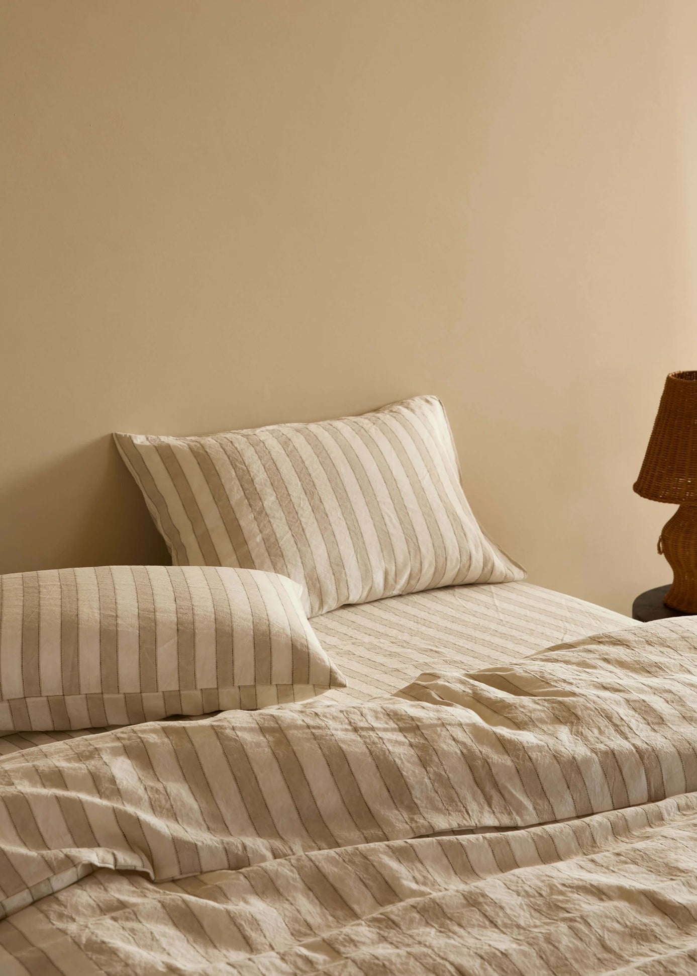 A neatly made bed with beige and cream striped bedding, including two pillows and a textured duvet, set against a warm beige wall with a dark wood table lamp on the right.