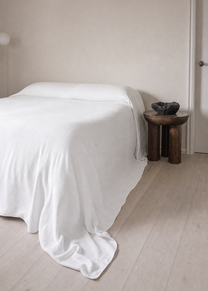 A minimalist bedroom scene featuring a bed dressed in crisp white bedding, positioned next to a small rustic wooden side table with a dark decorative bowl, set against a warm beige wall and light wood flooring.