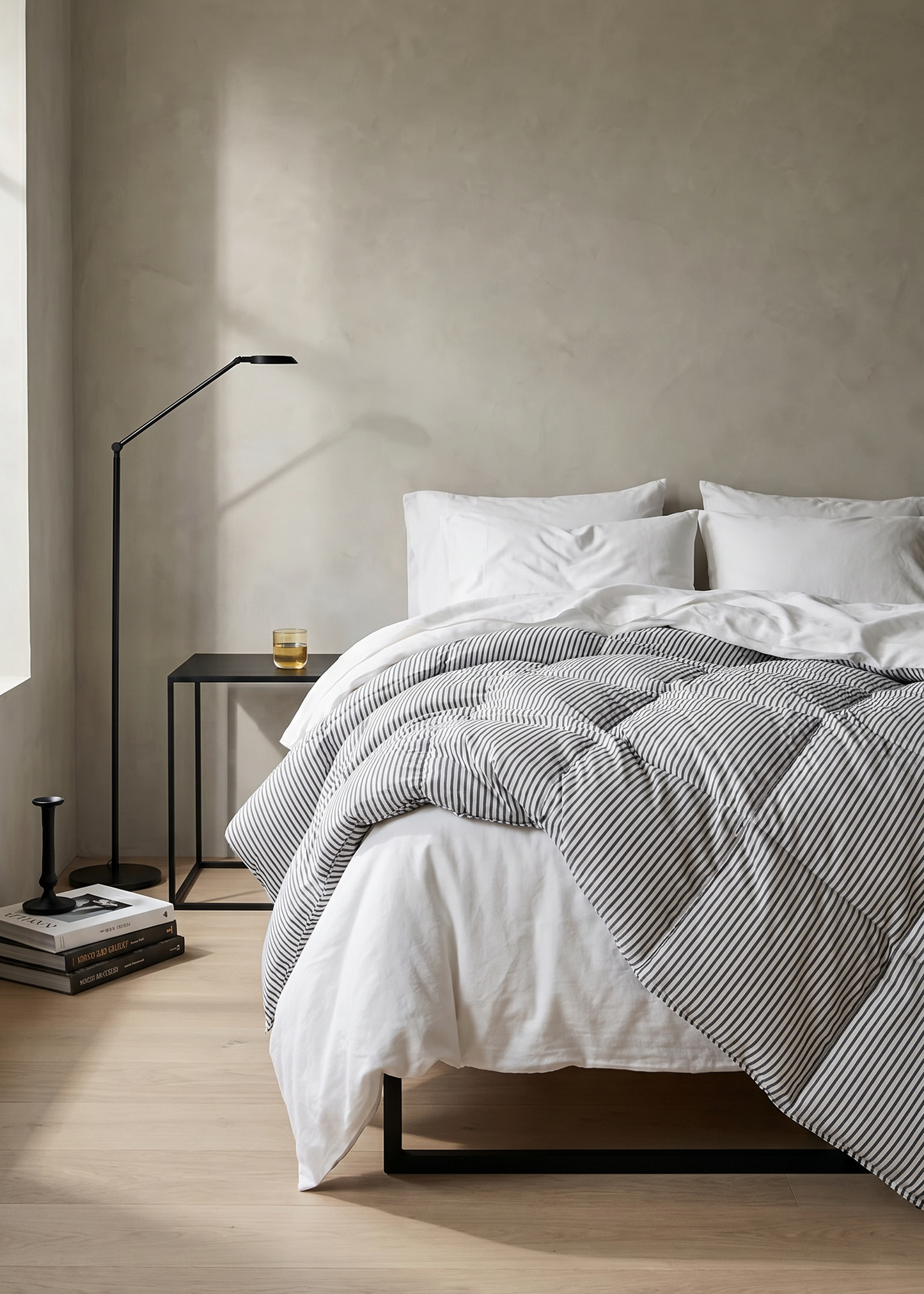 A minimalist bedroom scene featuring a bed dressed in white and gray striped bedding, set against a textured beige wall with a black floor lamp and side table.
