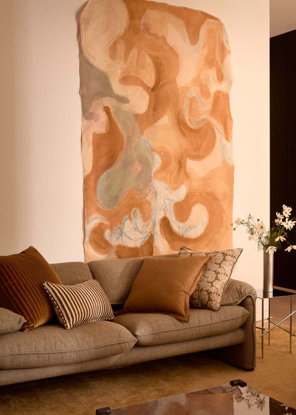 A large abstract wall art piece in warm peach, tan, cream, and muted blue-green tones hangs above a beige sofa in a styled living room with brown pillows, a glass coffee table, and a dark side cabinet.