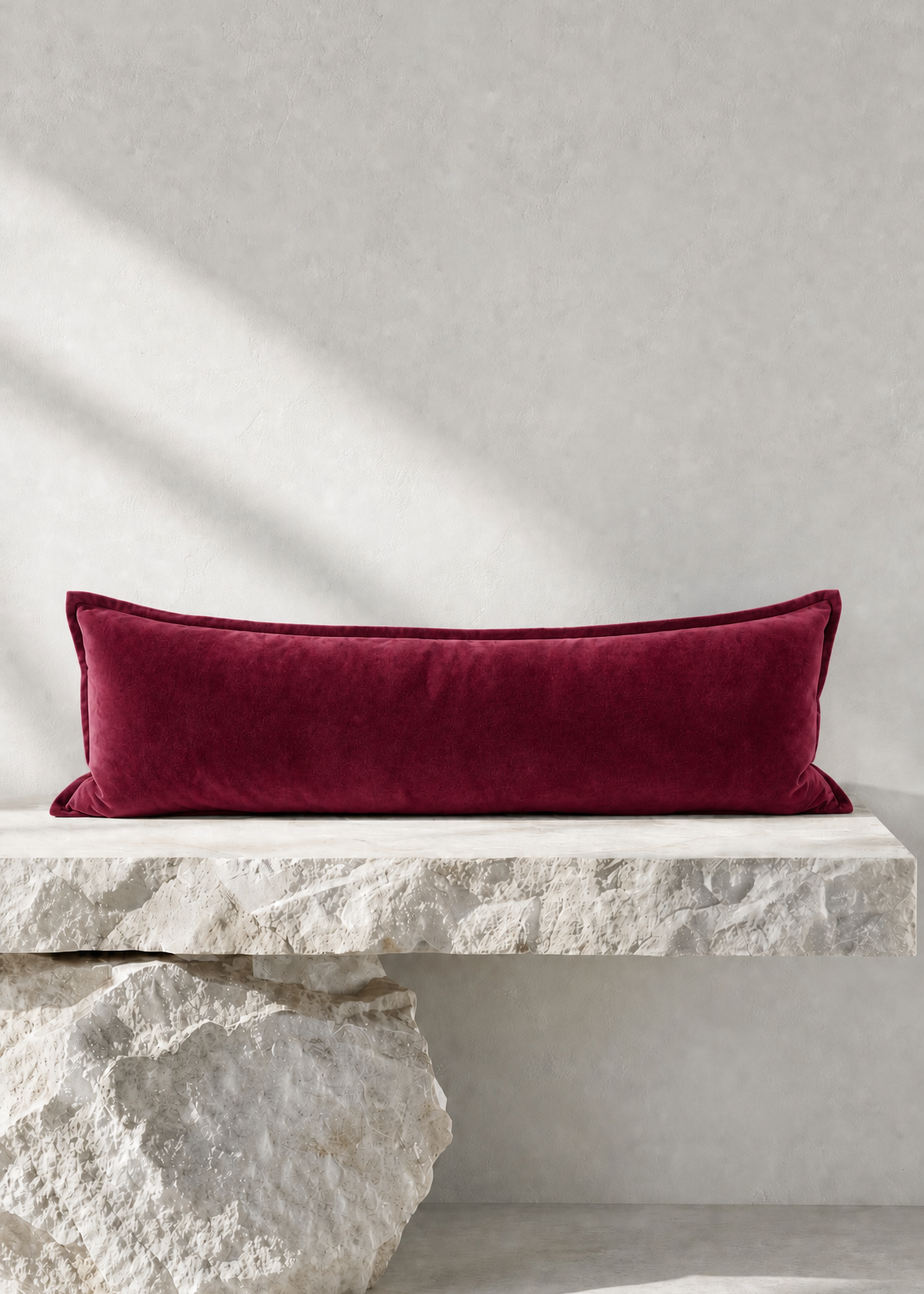 A rectangular deep burgundy velvet lumbar pillow displayed on a rustic white stone bench against a textured light gray wall.