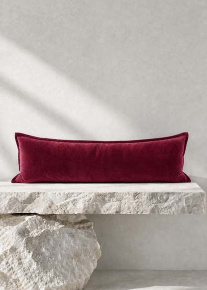 A rectangular deep burgundy velvet lumbar pillow displayed on a rustic white stone bench against a textured light gray wall.