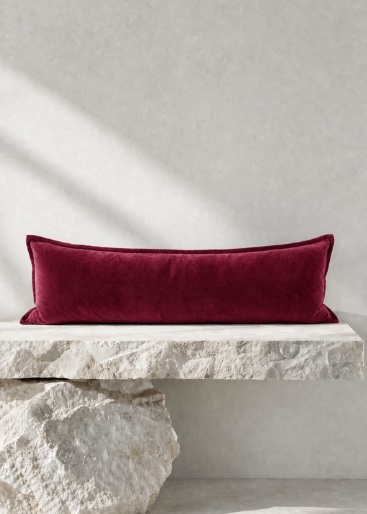 A rectangular deep burgundy velvet lumbar pillow displayed on a rustic white stone bench against a textured light gray wall.
